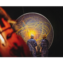 na zdjeciu widoczne dwie osoby stojące tyłem, w granatowych kombinezonach popisanych kolorowymi farbami, przed nimi czarna płaszczyzna w kształcie koła, na której narysowano kolorowe okręgi i elipsy