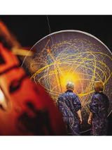 na zdjeciu widoczne dwie osoby stojące tyłem, w granatowych kombinezonach popisanych kolorowymi farbami, przed nimi czarna płaszczyzna w kształcie koła, na której narysowano kolorowe okręgi i elipsy