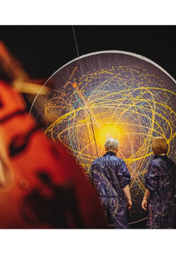 na zdjeciu widoczne dwie osoby stojące tyłem, w granatowych kombinezonach popisanych kolorowymi farbami, przed nimi czarna płaszczyzna w kształcie koła, na której narysowano kolorowe okręgi i elipsy