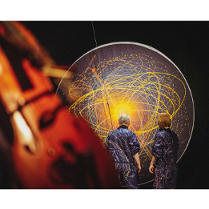 na zdjeciu widoczne dwie osoby stojące tyłem, w granatowych kombinezonach popisanych kolorowymi farbami, przed nimi czarna płaszczyzna w kształcie koła, na której narysowano kolorowe okręgi i elipsy