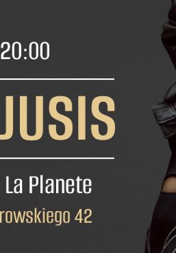 Zdjęcie przedstawia kobietę w kreatywnej, nietypowej pozie. Jej twarz jest ukryta za białymi krótkimi włosami. Ma na sobie skórzaną kurtkę i obcisły kombinezon.