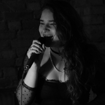 Zdjęcie, dziewczyna z mikrofonem w ciemnym pomieszczeniu. Dziewczyna ma ropuszczone długie włosy, koronkową bluzkę i naszyjnik z pereł.