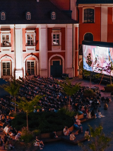 Pokaz filmowy na Wolnym Dziedzińcu. Na dziedzińcu Urzędu Miasta ogormny ekran kinowy, przed którym siedzą ludzie na leżakach i oglądają pokaz filmowy.