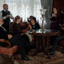 Zdjęcie, kadr filmu, aktorzy siedzą na eleganckich fotelach w salonie, w dłoniach trzymają filiżanki, przed nimi drewniany rzeźbiony stół.