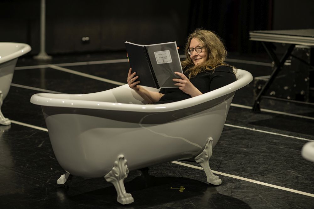 Kobieta czyta książkę leżąc w wannie będącej elementem scenicznej scenografii.