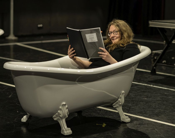 Kobieta leży w zdobionej, staroświeckiej wannie, która stoi na scenie jako rekwizyt. Uśmiecha się, czyta duży zeszyt.