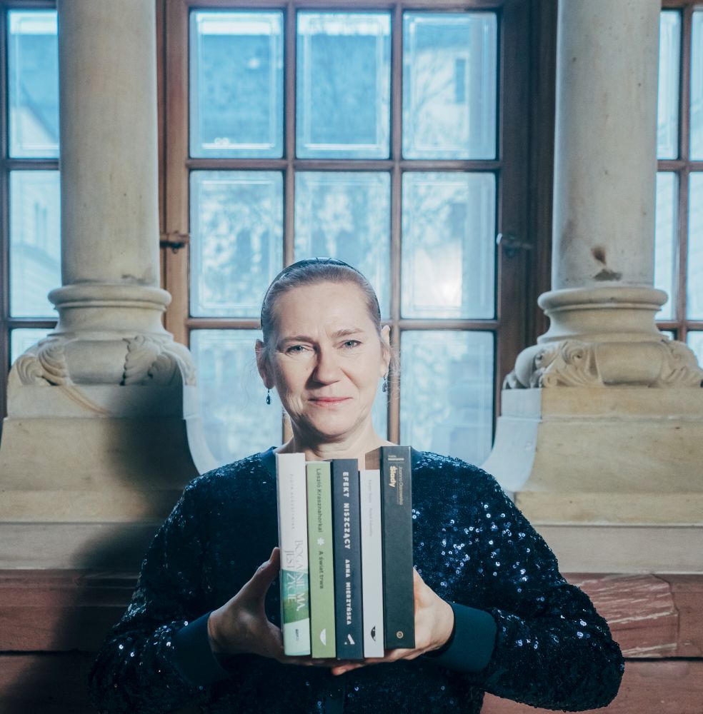 Kobieta prezentuje zestaw książek siedząc na tle okna.