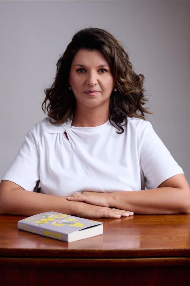 Kobieta w białej bluzce siedzi przy stole, na którym leży książka.