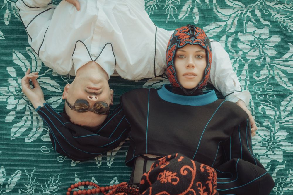 Kobieta w czepku i mężczyzna w okularach leżą na zielonym, wzorzystym dywanie.