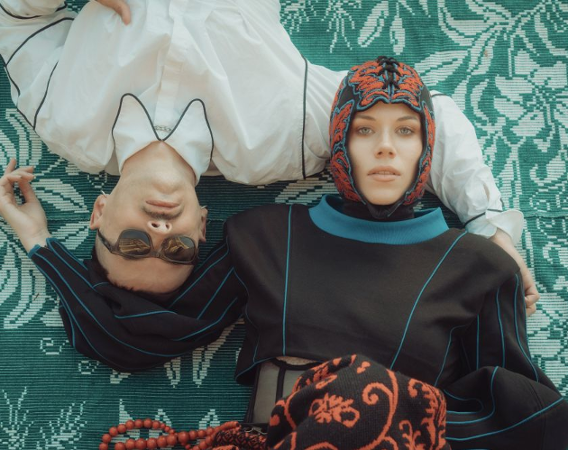 Kobieta w czepku i mężczyzna w okularach leżą na zielonym, wzorzystym dywanie.