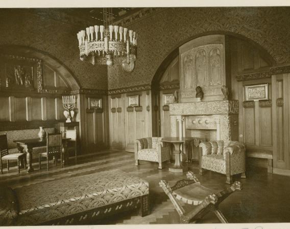 Komntaa zamkowa. Kominek w centrum. Obok stolik i dwa fotele. Duża ozdobna lampa i dywan. Drewniana boazeria.