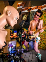 "Bajzel". Nagi gitarzysta przy mikrofonie. Gitara zakrywa jego brzuch, częśći intymne i uda. Z przodu nagi manekin kobiety z zaklejonymi taśmą piersiami.