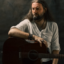 Mężczyzna w średnim wieku z długimi ciemnymi włosami i brodą. Ubrany w białą luźną koszulkę. Siedzi na wysokim krześle i jedną rękę opiera na gitarze akustycznej. Na ręce i na szyi ma biżuterie. Spogląda w bok.