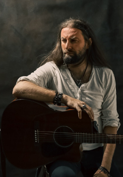Mężczyzna w średnim wieku z długimi ciemnymi włosami i brodą. Ubrany w białą luźną koszulkę. Siedzi na wysokim krześle i jedną rękę opiera na gitarze akustycznej. Na ręce i na szyi ma biżuterie. Spogląda w bok.