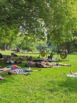 Zdjęcie, polana w parku, wokół drzewa, ludzie leżą na karimatach, przed nimi artyska gra na gongach i misach.