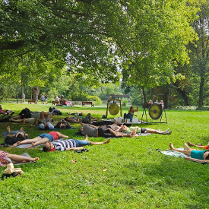Zdjęcie, polana w parku, wokół drzewa, ludzie leżą na karimatach, przed nimi artyska gra na gongach i misach.