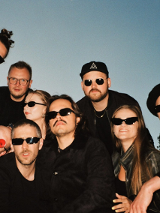 Zdjęcie grupy artystów stojących na tle nieba. Część osób kuca, część jest pochylona. Większość ma ciemne okulary. Wszyscy ubrani są w czarne ubrania.