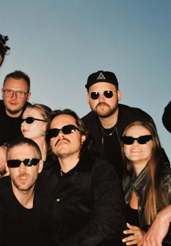 Zdjęcie grupy artystów stojących na tle nieba. Część osób kuca, część jest pochylona. Większość ma ciemne okulary. Wszyscy ubrani są w czarne ubrania.