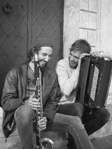 Dwóch śmiejących się mężczyzn siedzi u drzwi kamienicy, jeden trzyma akordeon, drugi saksofon.