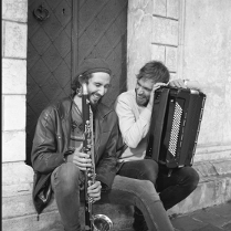 Dwóch śmiejących się mężczyzn siedzi u drzwi kamienicy, jeden trzyma akordeon, drugi saksofon.