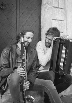 Dwóch śmiejących się mężczyzn siedzi u drzwi kamienicy, jeden trzyma akordeon, drugi saksofon.