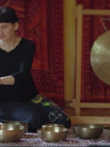 Zdjęcie przedstawia siedzącą kobietę grającą na misie. Na tle ściany pokrytej czerwoną tkaniną w azjatyckie wzory stoi gong.