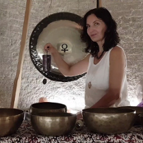 Zdjęcie przedstawia kobietę siedzącą przed dużym gongiem oraz kilkoma misami do śpiewu. Kobieta trzyma mały, czarny, prostokątny przedmiot w swojej prawej ręce. Ma na sobie białą koszulkę bez rękawów i naszyjnik. Gong jest zawieszony na suficie za kobietą. Misy do śpiewu są ułożone w półkolu przed nią. Tło stanowi biała ściana. Kobieta ma ciemne włosy i patrzy w obiektyw aparatu. Zdjęcie wydaje się być wykonane w pomieszczeniu z białą ceglaną ścianą. Kobieta lekko się uśmiecha.