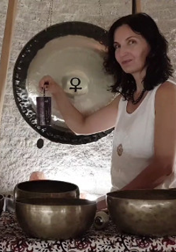 Zdjęcie przedstawia kobietę siedzącą przed dużym gongiem oraz kilkoma misami do śpiewu. Kobieta trzyma mały, czarny, prostokątny przedmiot w swojej prawej ręce. Ma na sobie białą koszulkę bez rękawów i naszyjnik. Gong jest zawieszony na suficie za kobietą. Misy do śpiewu są ułożone w półkolu przed nią. Tło stanowi biała ściana. Kobieta ma ciemne włosy i patrzy w obiektyw aparatu. Zdjęcie wydaje się być wykonane w pomieszczeniu z białą ceglaną ścianą. Kobieta lekko się uśmiecha.