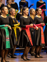 Chór dziewczęcy w czarnych sukienkach przepasanych kolorowymi wstęgami.