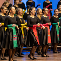 Chór dziewczęcy w czarnych sukienkach przepasanych kolorowymi wstęgami.