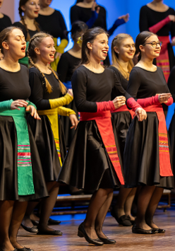 Chór dziewczęcy w czarnych sukienkach przepasanych kolorowymi wstęgami.