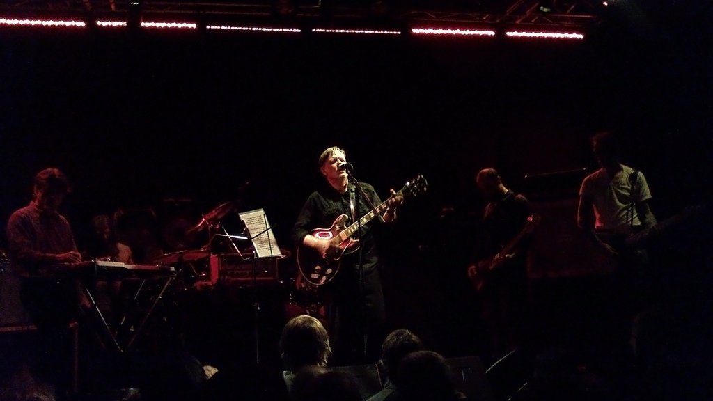 Oświetlony reflektorem gitarzysta zespołu rockowego Swans w ciemnej sali koncertowej, za nim, w półmroku pozostali członkowie zespołu: po lewej klawiszowiec, po prawej dwaj gitarzyści, perkusista w głębi kadru.