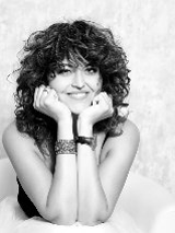 Czarno-białe zdjęcie przedstawia siedzącą i uśmiechającą się kobietę, która opiera swoją głowę na rękach wspartych łokciami o kolana.