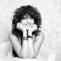 Czarno-białe zdjęcie przedstawia siedzącą i uśmiechającą się kobietę, która opiera swoją głowę na rękach wspartych łokciami o kolana.