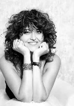 Czarno-białe zdjęcie przedstawia siedzącą i uśmiechającą się kobietę, która opiera swoją głowę na rękach wspartych łokciami o kolana.