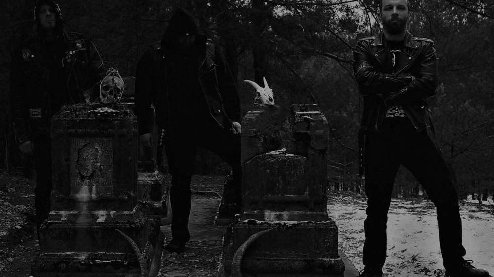 Ciemna fotografia w czarno-białych kolorach, trzech ubranych na czarno mężczyzn stoi w otoczeniu nagrobków, na których spoczywają przedmioty przypominające zwierzęce czaszki.