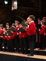 Grupa chłopców ubranych w czarne spodnie, białe koszule i czerwone swetry. W rękach trzymają duże zeszyty, w ktorych są nuty.