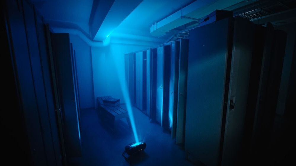 Kadr z filmu - pomieszczenie przypomina szatnię lub przebieralnię na siłowni, jest w nim ciemno, na podłodze leży urządzenie, z którego bije wąski, błękitny promień światła w kierunku sufitu.