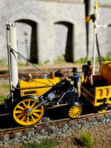 To jest miniaturowy model parowozu Rocket, historycznego pociągu, który zrewolucjonizował transport. Został ustawiony na makiecie torów kolejowych, kompletny z miniaturowym wagonem węglowym i figurkami ludzi. Model jest bardzo szczegółowy, z dokładnymi reprezentacjami projektu lokomotywy, w tym kotła, kół i nawet kominu. Model jest również pomalowany w jaskrawych kolorach, co dodaje realizmu.