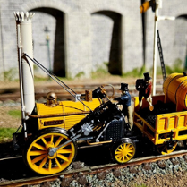 To jest miniaturowy model parowozu Rocket, historycznego pociągu, który zrewolucjonizował transport. Został ustawiony na makiecie torów kolejowych, kompletny z miniaturowym wagonem węglowym i figurkami ludzi. Model jest bardzo szczegółowy, z dokładnymi reprezentacjami projektu lokomotywy, w tym kotła, kół i nawet kominu. Model jest również pomalowany w jaskrawych kolorach, co dodaje realizmu.