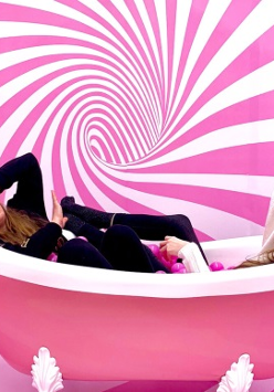 Dwie dziewczyny w ubraniu i w czapkach w różowej wannie. Na ścianie biało-różowy wir. W dłoniach maja różowe kulki. Dziewczyna po lewej ma wyprostowana do góry rękę.