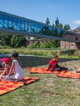 Grupa dzieci leży na kocach na trawniku. W tle widać szklaną kładkę prowadzącą nad rzeką do starego, ceglanego budynku.