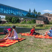 Grupa dzieci leży na kocach na trawniku. W tle widać szklaną kładkę prowadzącą nad rzeką do starego, ceglanego budynku.