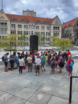 Zdjęcie, grupa osób stoi przed pomnikiem, za nimi ulica, drzewa, dwa samochody.