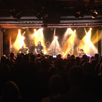 Na pierwszym planie głowy widowni, która słucha koncertu. Na oświetlonej scenie piątka mężczyzn w średnim wieku gra koncert na różnych instrumentach. Wśród nich jest gitarzysta, bębniarz, wiolonczelista, akordeonista.