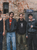 Czworo młodych mężczyzn stoi obok siebie na tle starego, ceglano-kamiennego muru zniszczonego budynku. Po prawej stronie widoczne są drewniane drzwi z tabliczką "Ładna".