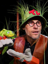 Zdjęcie aktora w zielonym kapeluszu, porośniętym trawami, który trzyma w ręce pacynkę żabę.