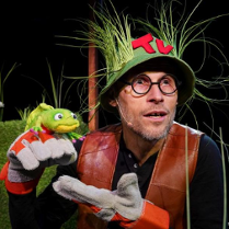 Zdjęcie aktora w zielonym kapeluszu, porośniętym trawami, który trzyma w ręce pacynkę żabę.