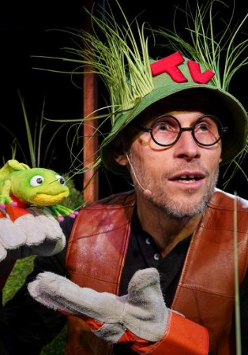 Zdjęcie aktora w zielonym kapeluszu, porośniętym trawami, który trzyma w ręce pacynkę żabę.