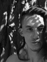 Czarno-białe zdjęcie przedstawia twarz tancerza Patryka Jarczoka, patrzącego na wprost w kierunku, z którego wykonano zdjęcie. Tancerz znajduje się w cieniu.